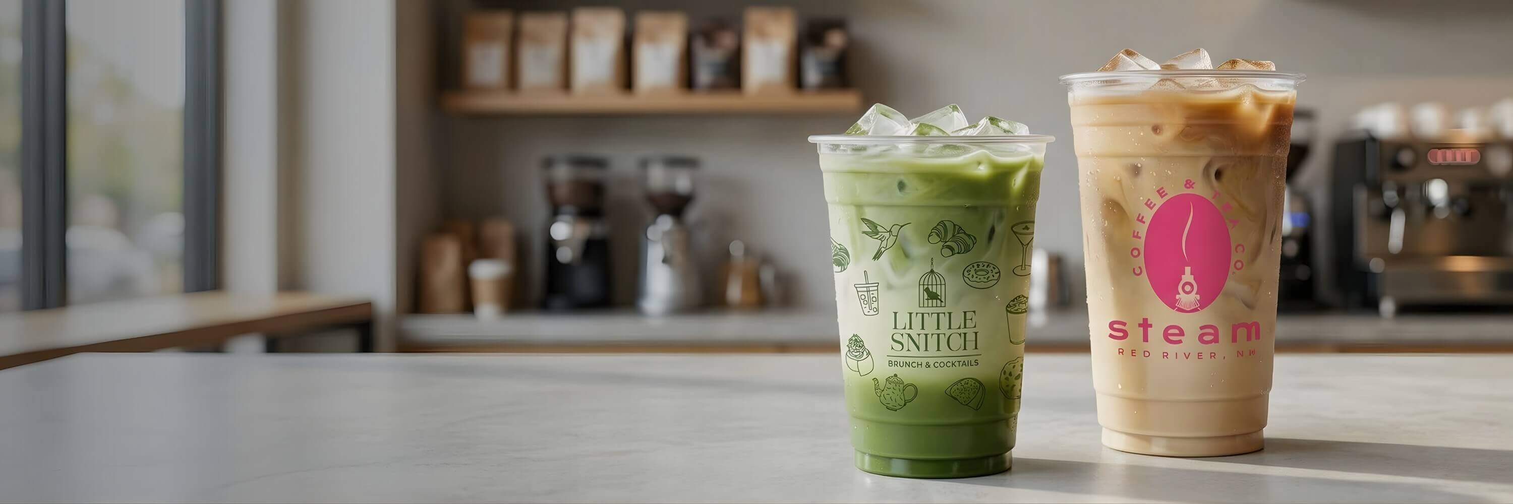 Two custom-printed clear PET plastic cups on a cafe counter, one featuring "Little Snitch" green branding with an iced matcha and the other "Steam" pink branding with an iced latte.
